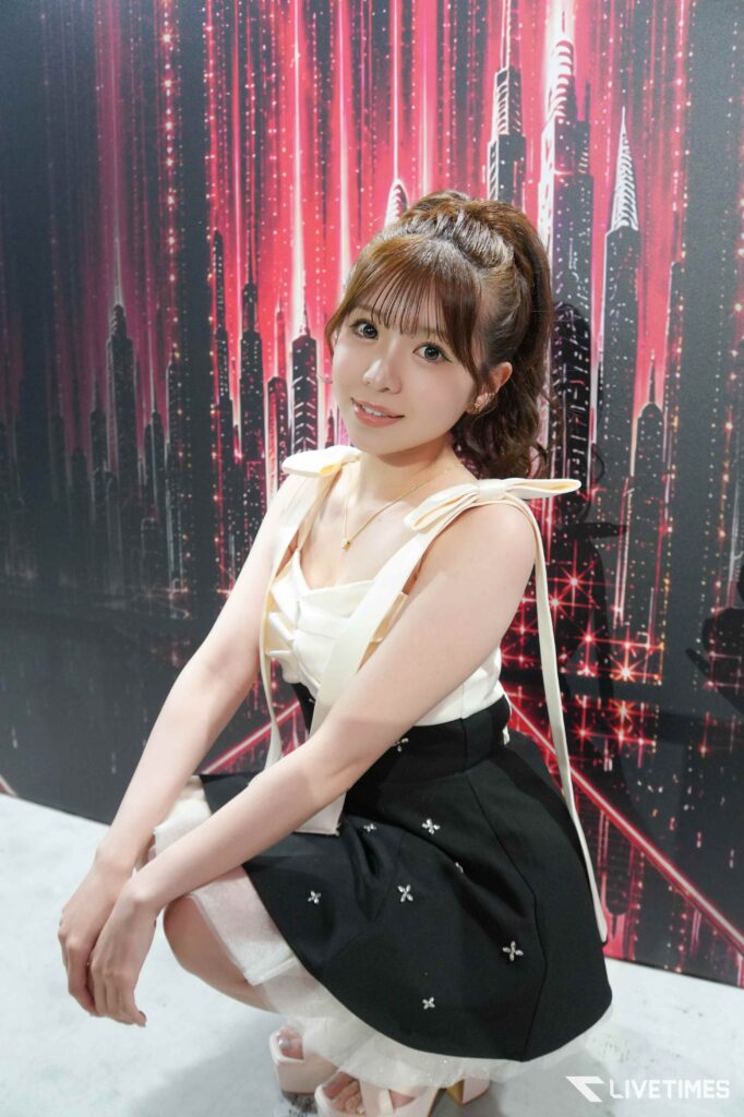 Young woman with brown hair in a high ponytail, smiling, wearing a white top with ribbons and a black skirt, posed in front of a pink neon cityscape backdrop.