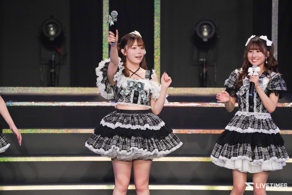 Two female performers in matching black-and-white plaid costumes on a glittery stage, one raising a microphone.