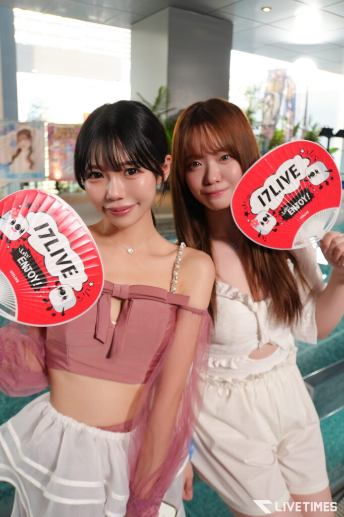 Two young women posing for a photo, each holding a red IZLIVE fan with white text, wearing pink and white outfits indoors at an event.