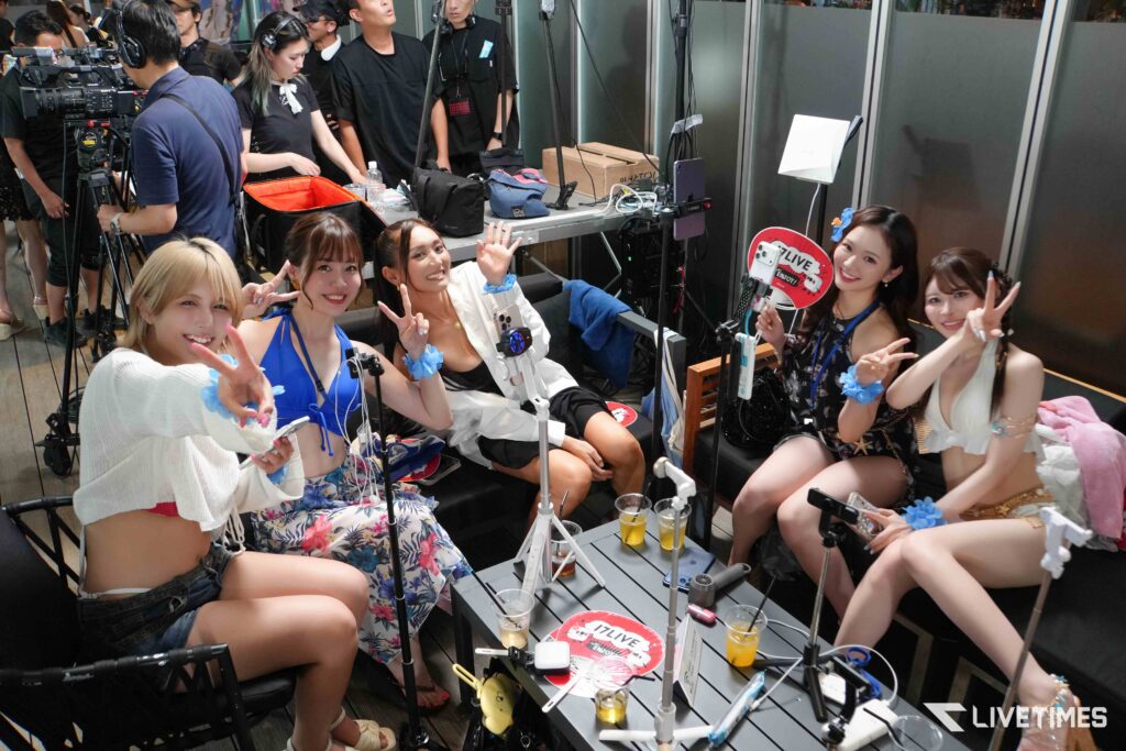 Backstage group photo of several young women posing and smiling, with cameras and crew in the background.