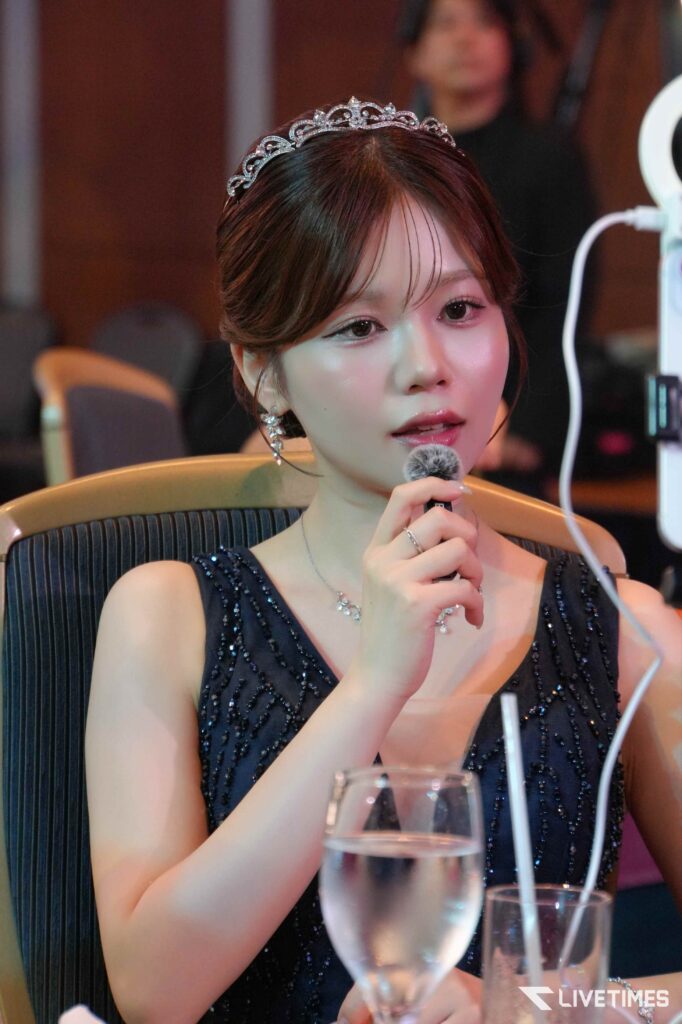 Woman wearing a jeweled tiara and beaded evening gown speaking into a microphone at a formal event, with glassware on the table in front of her.