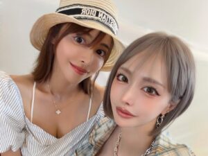Two friends posing a close selfie, one wearing a straw hat and striped top, the other in a plaid shirt with gold jewelry, both smiling at the camera.