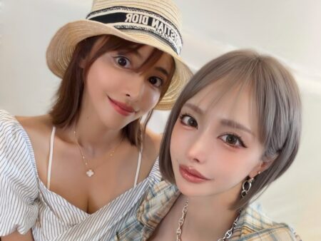 Two friends posing a close selfie, one wearing a straw hat and striped top, the other in a plaid shirt with gold jewelry, both smiling at the camera.