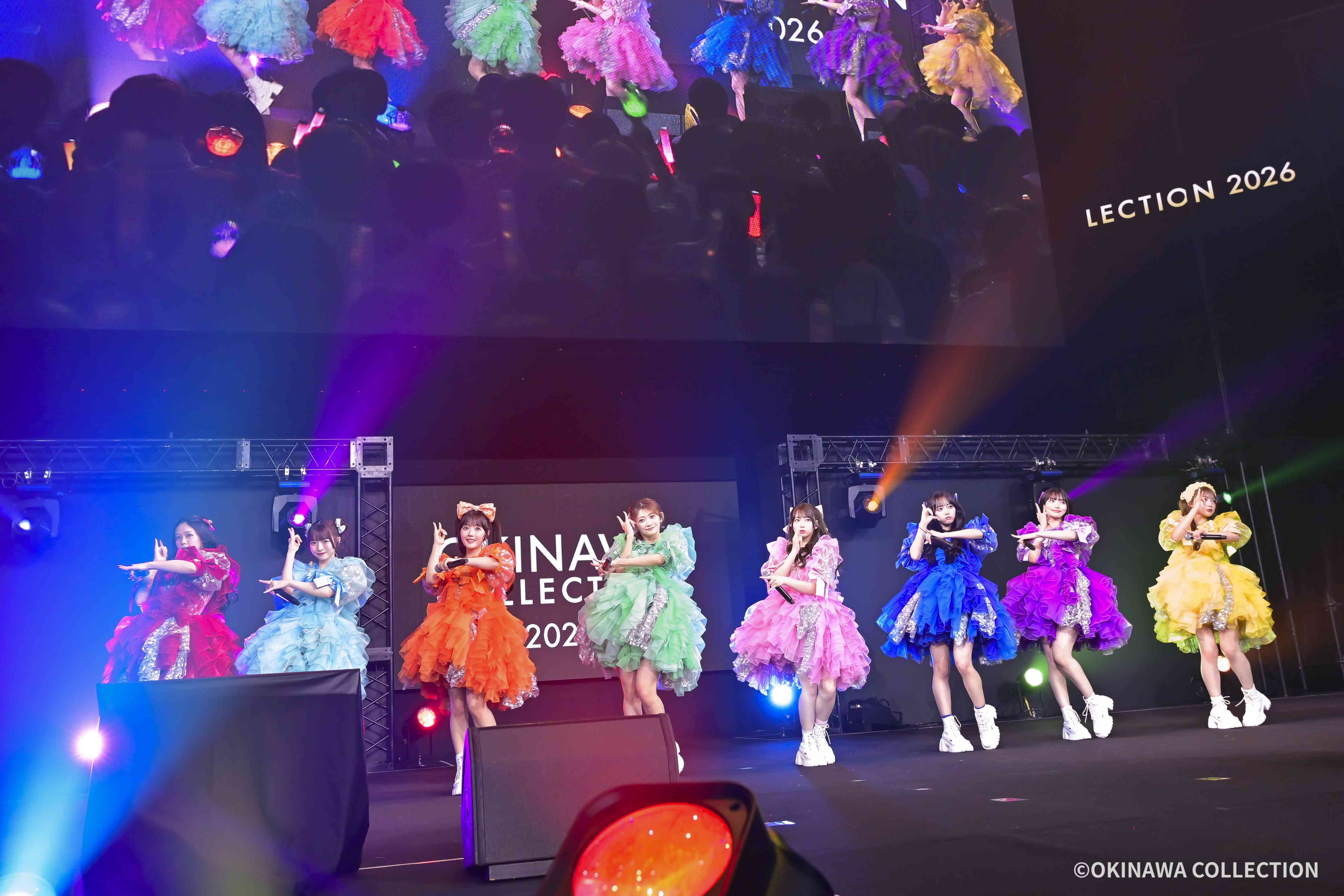 Group of performers in bright, ruffled dresses dancing on a stage with colorful lighting and an audience in the background. The banner shows 'OKINAWA COLLECTION'.