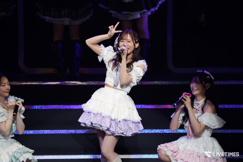 Female pop group performing on stage in frilly white outfits; central singer poses with peace sign and microphone under purple lights.