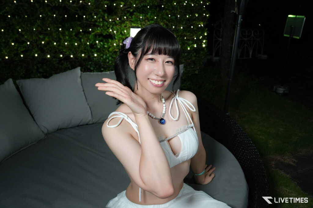 Smiling young woman in a white dress waving outdoors at night, with string lights in the background and jewelry on.