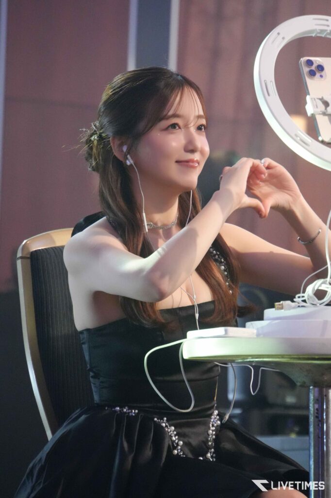 Smiling woman in a black dress forms a heart shape with her hands beside a ring light.