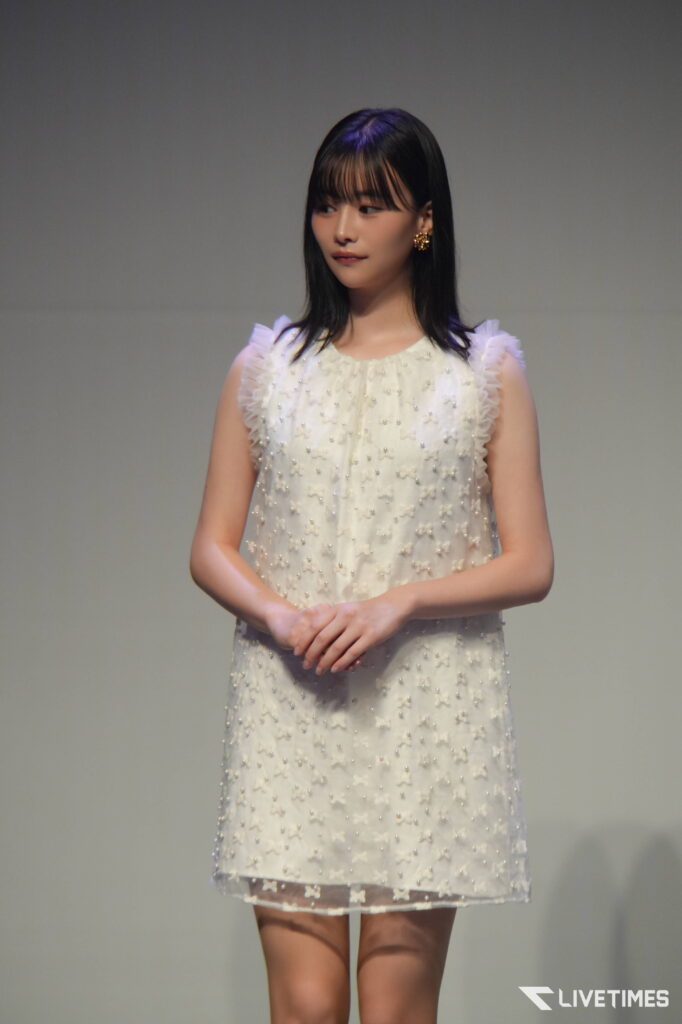 Young woman wearing a white beaded dress standing on a stage, hands clasped in front of her, neutral backdrop behind her