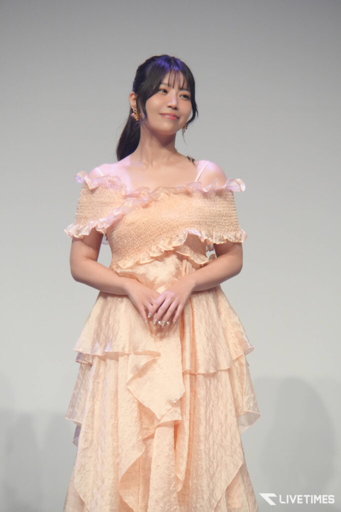 Young woman in a peach off-shoulder dress on stage, hands clasped, smiling softly.