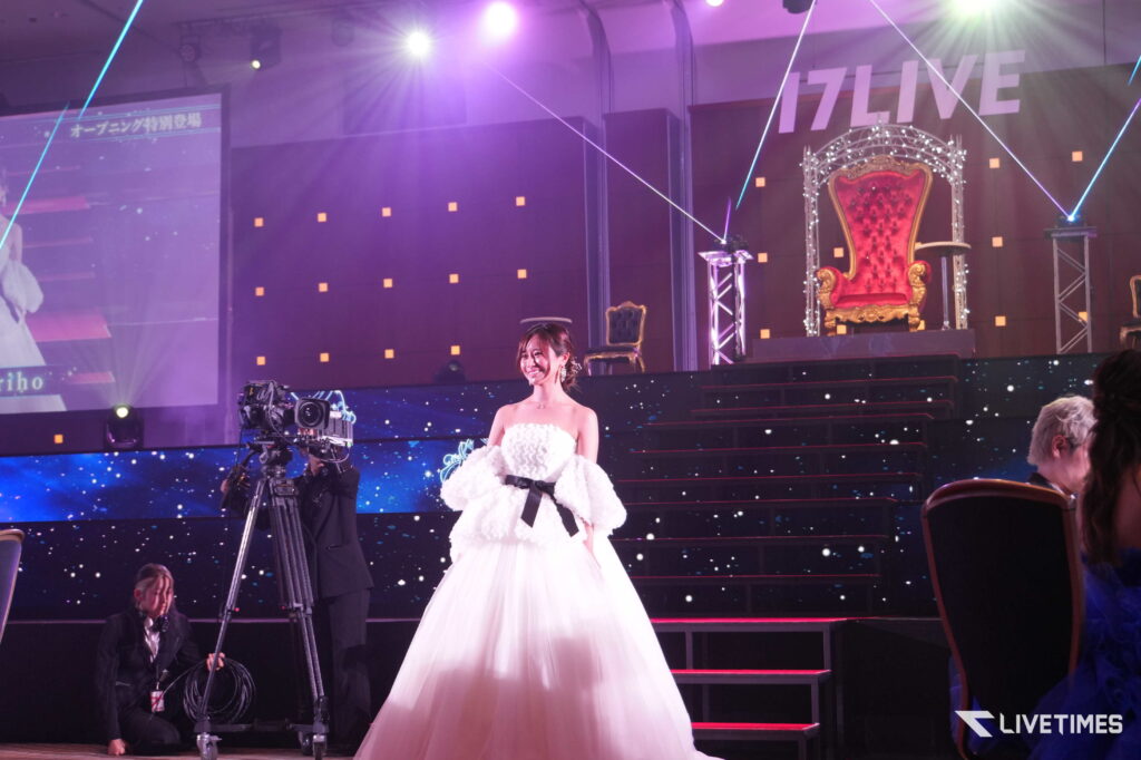 A woman in a white formal gown stands on a lit stage with a large ornate red throne to the right and a camera on a tripod filming from the left, purple lighting overhead.
