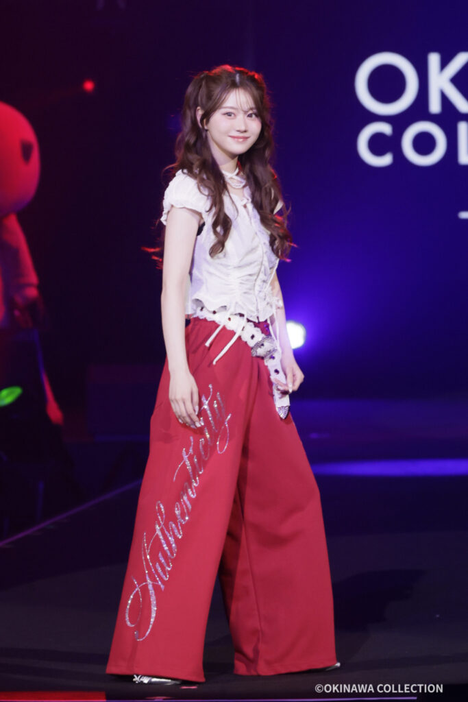 Young woman in a white blouse and red wide-leg pants posing on a dimly lit stage with purple lighting.