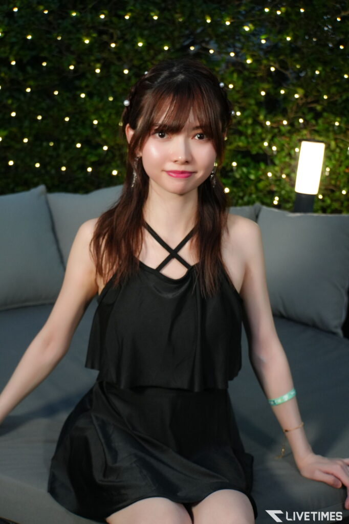 Young woman in a black halter dress seated on a couch, with string lights and greenery in the background.