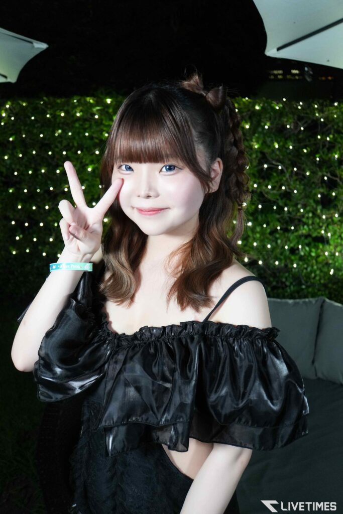 Young woman making a peace sign in front of a wall of fairy lights, wearing a shiny black off‑the‑shoulder dress.