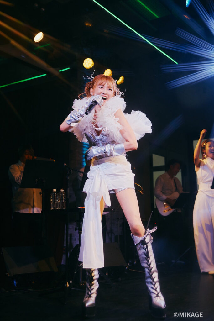 Female singer in a white ruffled costume and silver knee-high boots performs on stage, microphone in hand with a live band in the background and colorful lights.