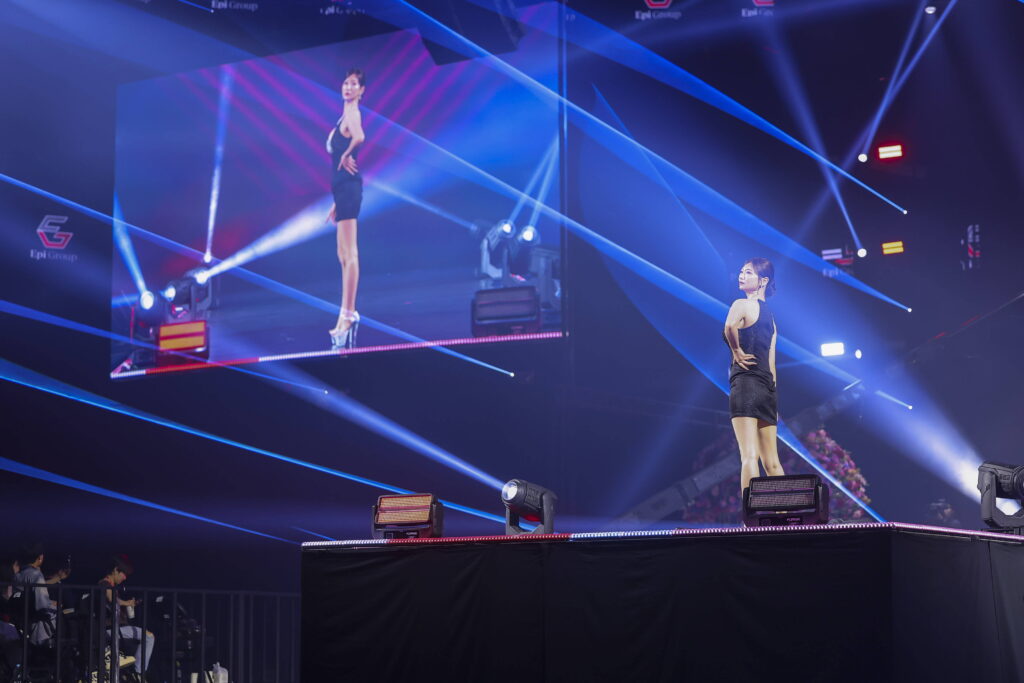 Two models in black cocktail dresses pose on a brightly lit stage with blue laser lighting during a fashion show.