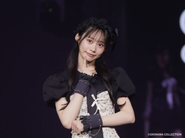 Young female performer on a dark stage, posing with a finger to her chin in a black puff-sleeve top and cream polka-dot dress with a bow.