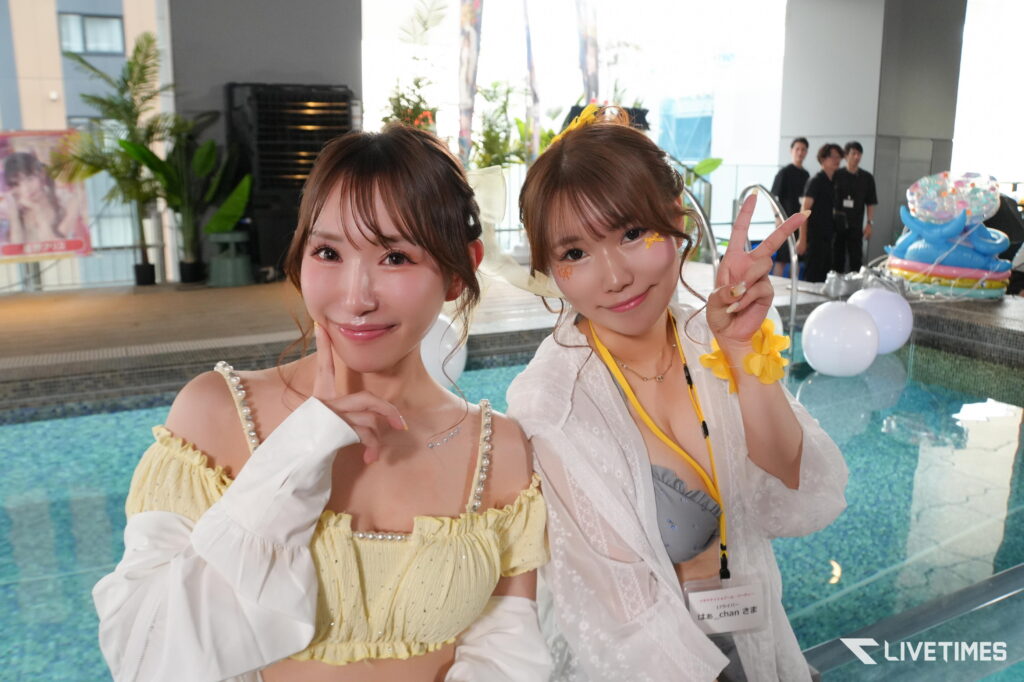 Two young women pose by a pool at a party, wearing yellow and white outfits with accessories and festive face decorations visible in the background.