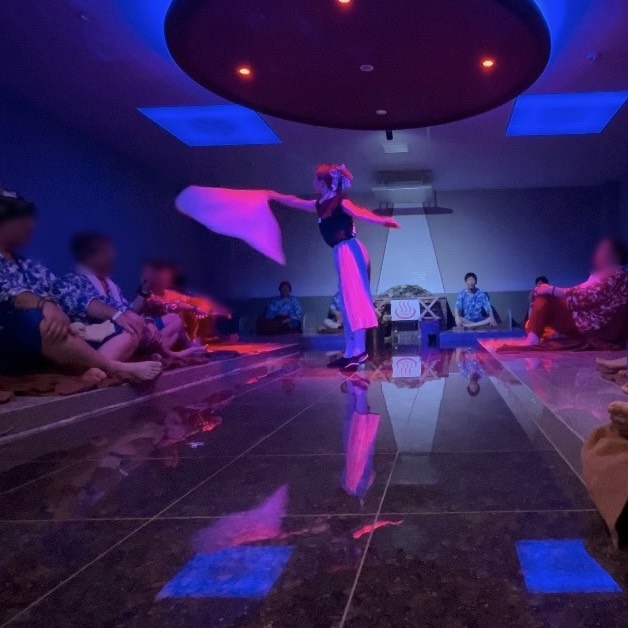Dancer in blue-white outfit twirls with a pink scarf in a blue-lit room, audience seated on the floor around a glossy stage area.