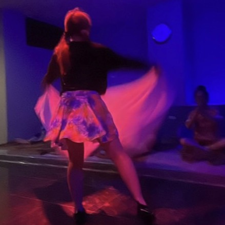 Dancer seen from behind performing on a stage with motion blur; wearing a floral skirt under blue lighting.