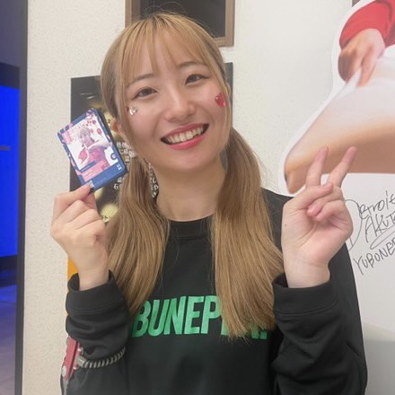 Young woman with blond hair smiling, holding a small rectangular card in her left hand and flashing a peace sign with her right hand indoors.