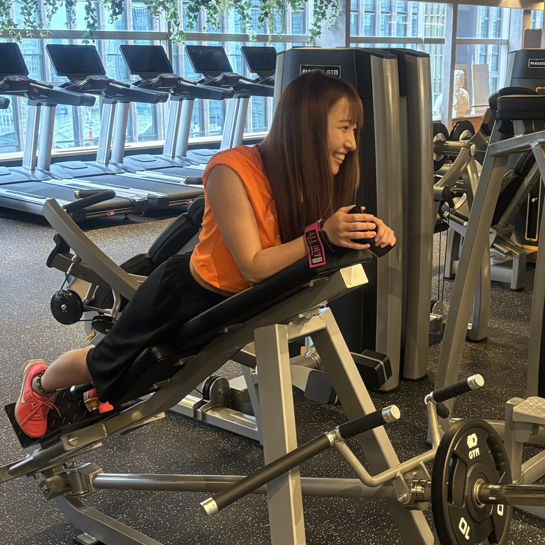 Woman in an orange shirt sits on a leg-extension machine at the gym, smiling with hands resting on the pad. Parked treadmills and gym equipment visible in the background.