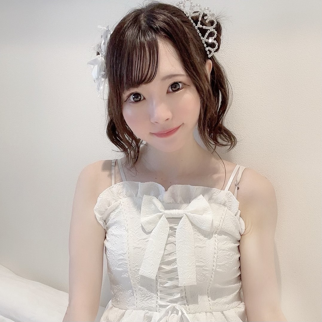 Young woman in a white lace dress with a large bow, wearing a tiara and white hair accessories, smiling at the camera against a pale wall.