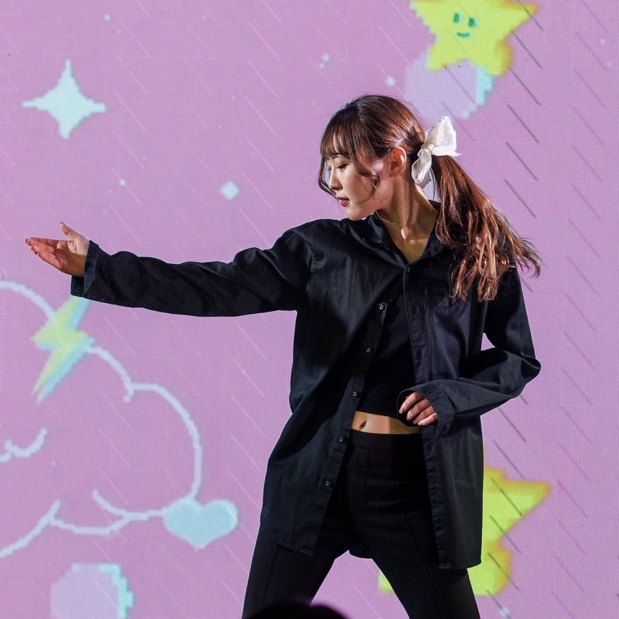 Female dancer in a black oversized shirt and leggings performs a pose on stage, with a pink starry backdrop.