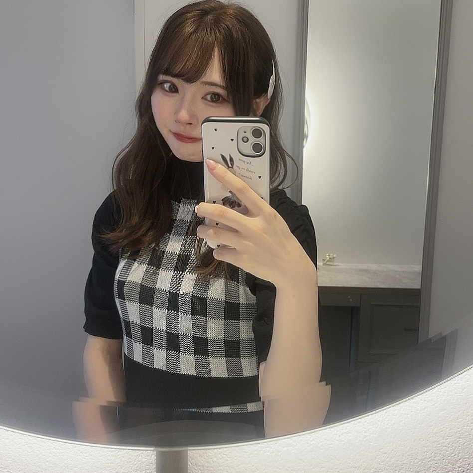 Woman taking a mirror selfie with a smartphone, wearing a black-and-white checkered top and a hair clip in a softly lit bathroom.