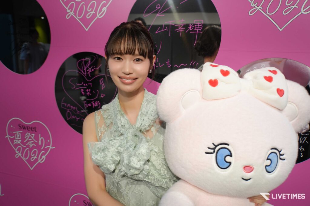Young woman in a lace mint dress smiles beside a large white stuffed bear with red heart accents against a pink backdrop.