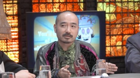 Bearded man in a patterned jacket speaks during a panel discussion in a studio setting with a colorful backdrop behind him.