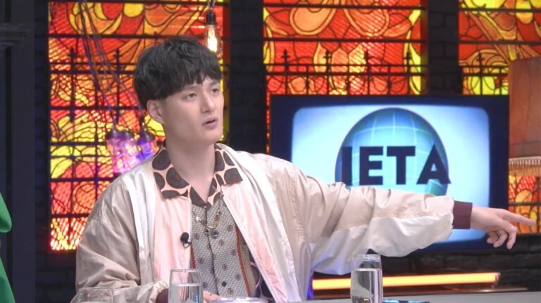 Host in a light jacket gestures to the right while seated at a colorful TV set with a stained-glass backdrop and a screen showing 'ETA' in the background.