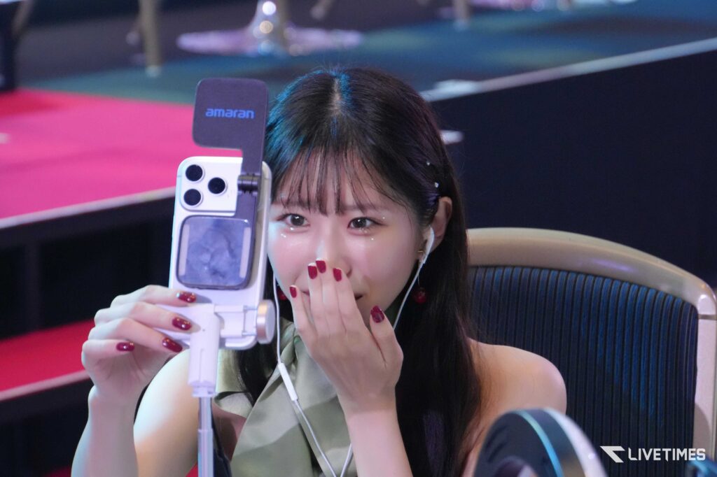Woman smiling and taking a selfie with a phone on a selfie stick, wearing AirPods, in a colorful event setting.