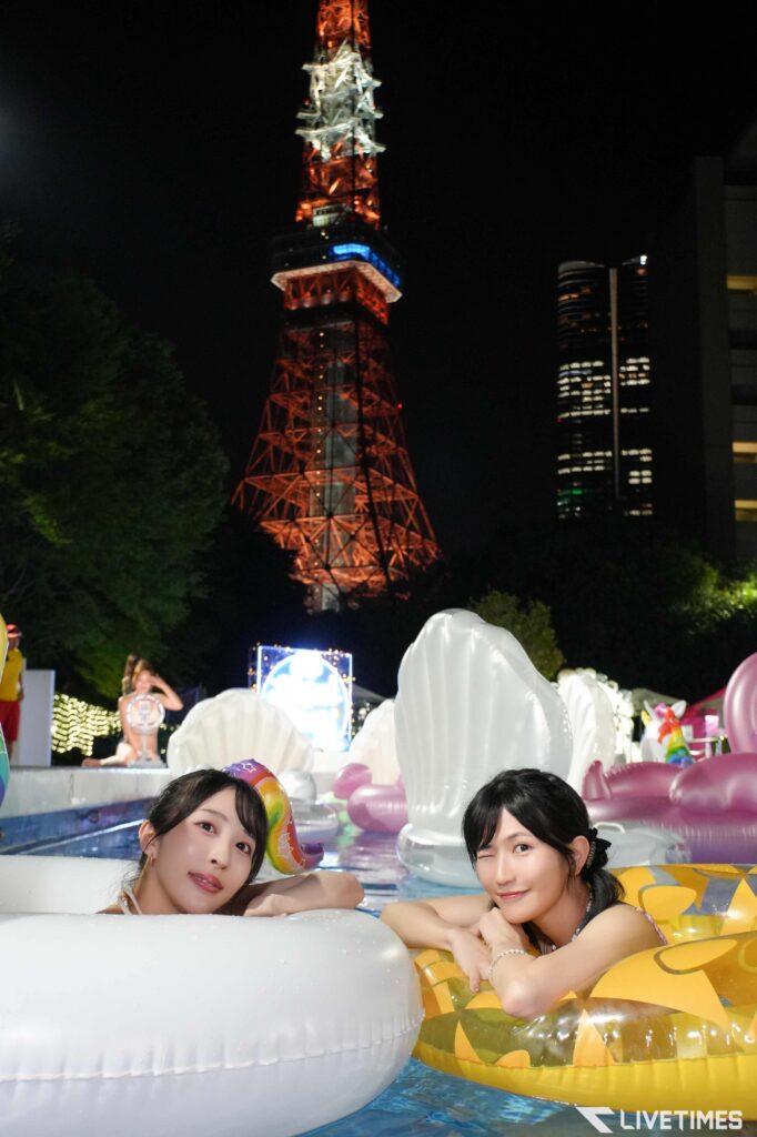 Nighttime scene at a pool party with two women in inner tubes; Tokyo Tower lit orange in the background.