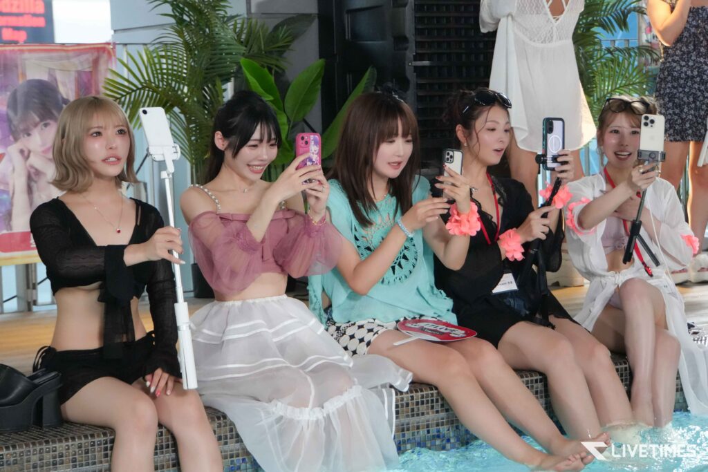 Group of young women sitting by a pool, taking selfies with their smartphones; colorful outfits and leis, tropical plants in the background.