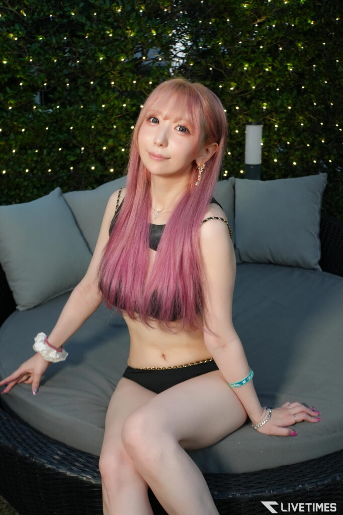 Young woman with long pink hair sits on a gray outdoor couch, smiling, with string lights and greenery behind her.