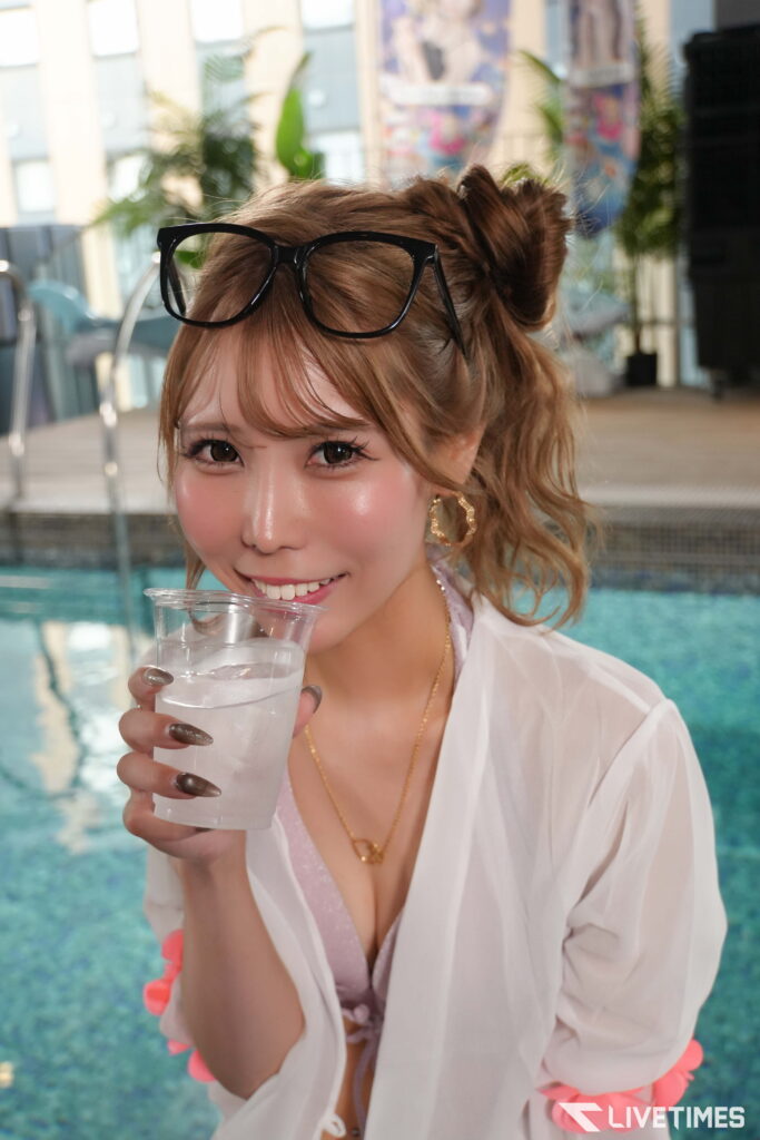 Smiling woman by a pool with black sunglasses on her head, holding a plastic cup of water.
