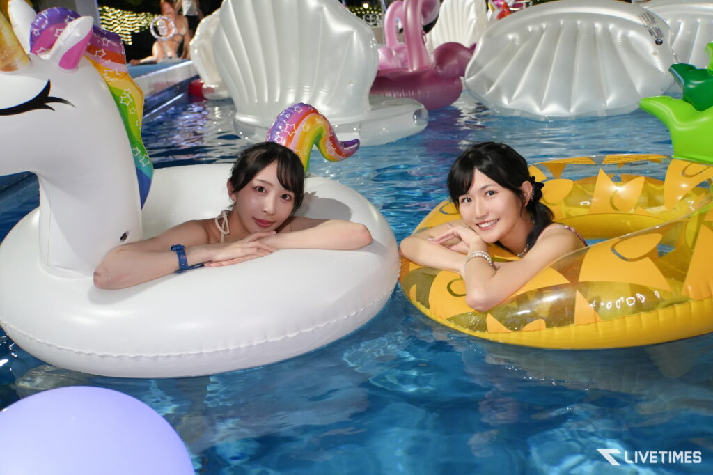 Two young women lean on oversized inflatable pool floats—a white unicorn with a rainbow horn and a yellow pineapple—in a bright pool setting.