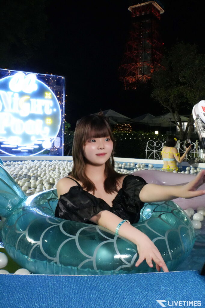 Woman reclining on a teal mermaid-scale pool float at night, with string lights and a red-lit tower in the background.