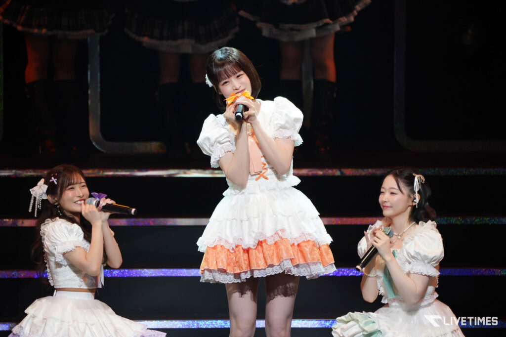 Center-stage female singer in a white lace dress smiling into an orange-bowed microphone, flanked by two other performers in matching outfits on a concert stage.