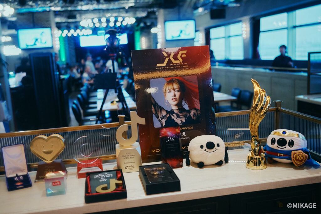 Table full of awards and memorabilia in a blue-lit studio, foreground includes a panda plush and a gold trophy.