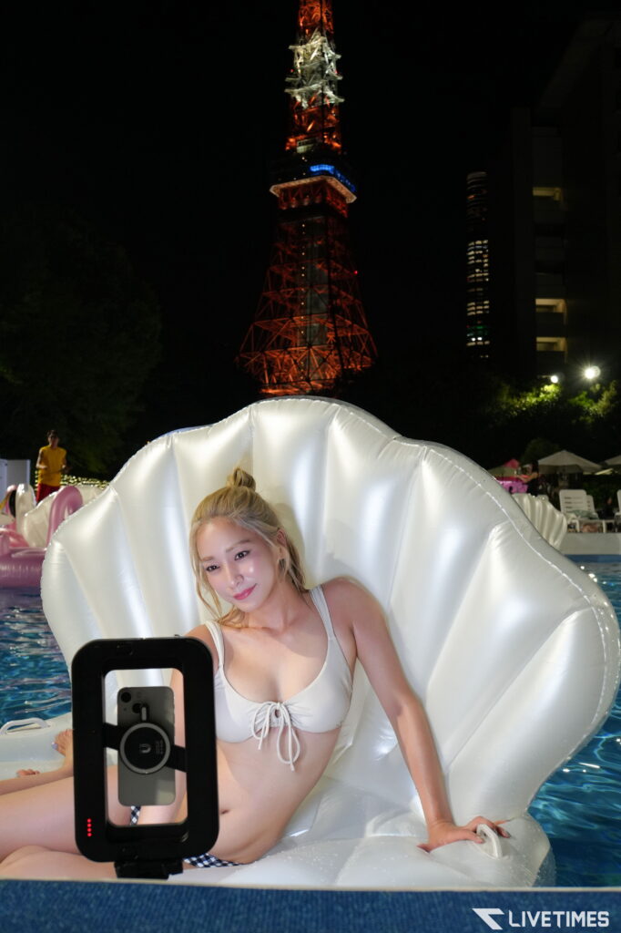Woman in a white bikini on a large white shell-shaped inflatable, taking a selfie with her phone at night, Tokyo Tower lit red in the background.