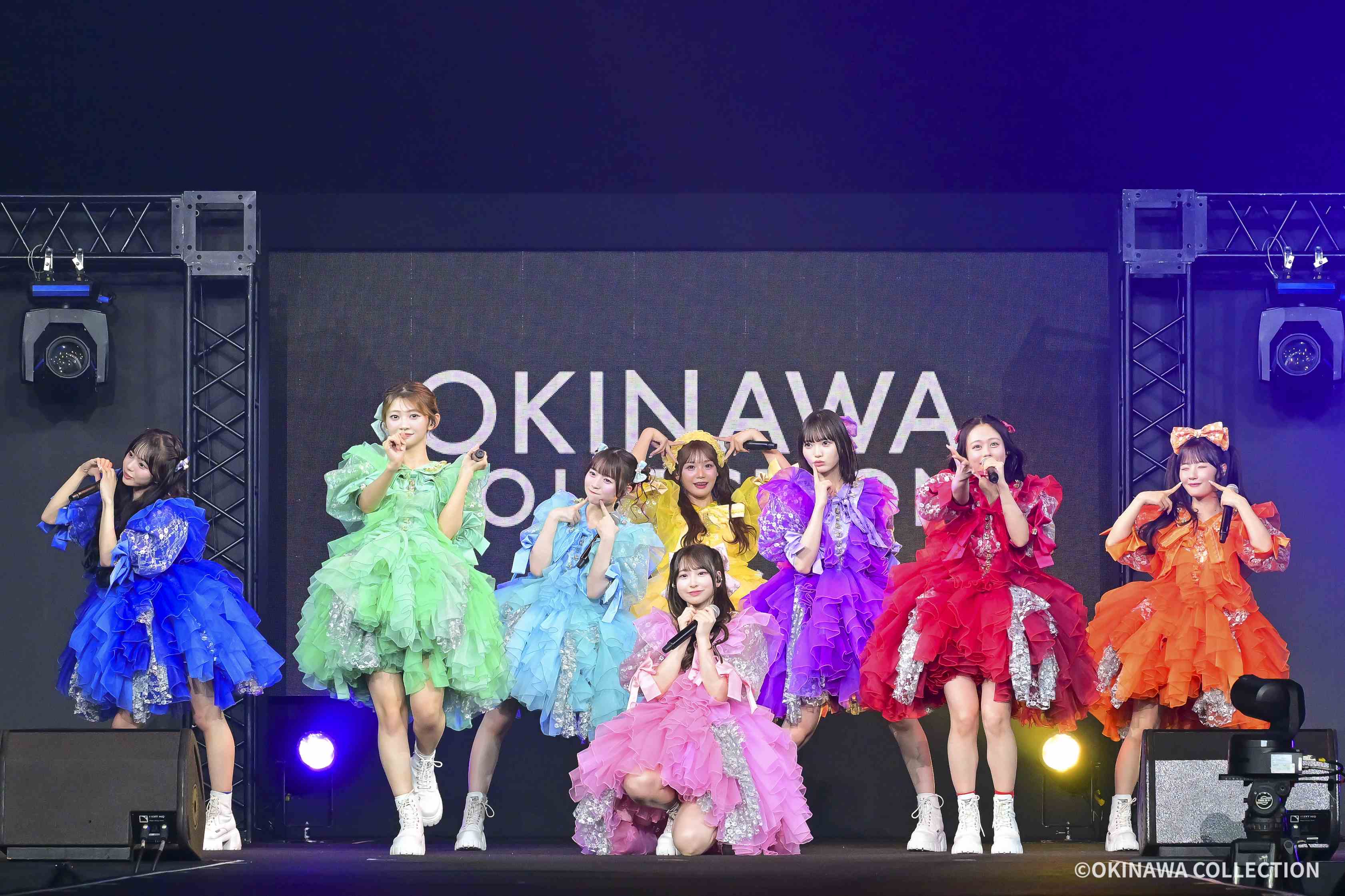 Group of performers in bright, frilly dresses (blue, green, yellow, pink, purple, orange) singing into microphones on a stage with a large screen behind them and stage lights above.