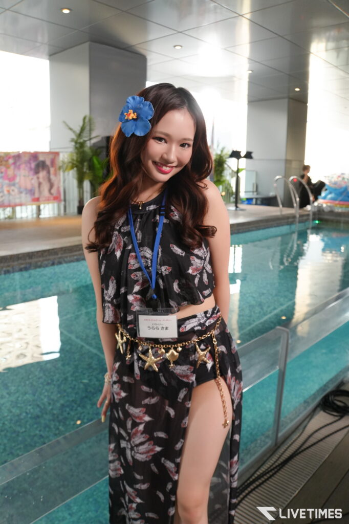 Smiling young woman by an indoor pool with a blue hibiscus in her hair and a black floral outfit, facing the camera.