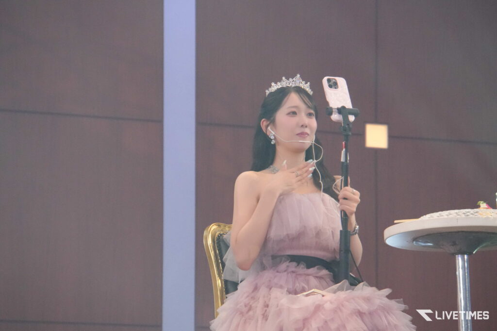 Young woman in a pink gown wearing a tiara and microphone headset, posing on stage with a smartphone on a stand beside her.