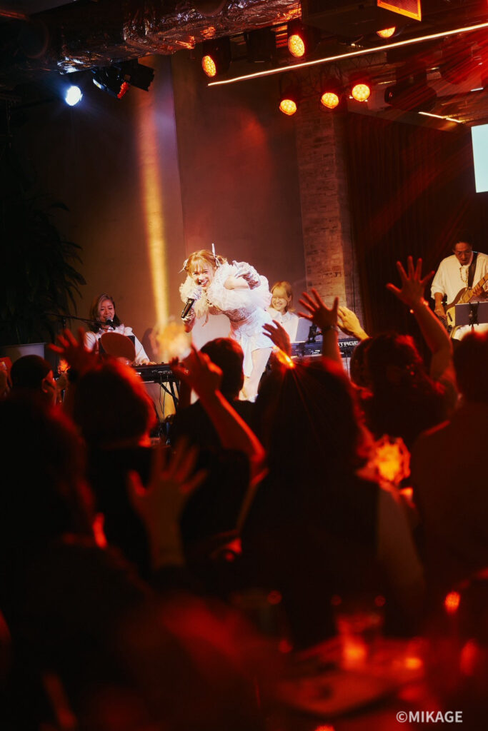 Singer in white dress performs on stage with a live band, microphone in hand, as a cheering crowd raises their hands amid red stage lights.