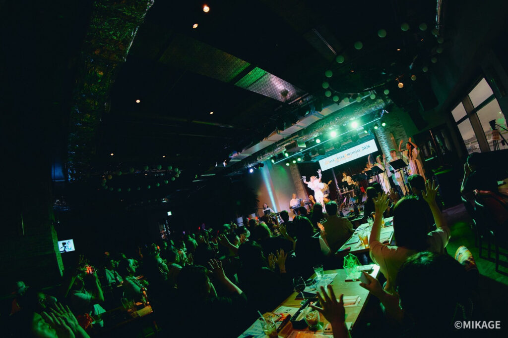 Crowd raises hands and cheers at a live band on a green-lit stage in a dark club, tables with drinks in the foreground.