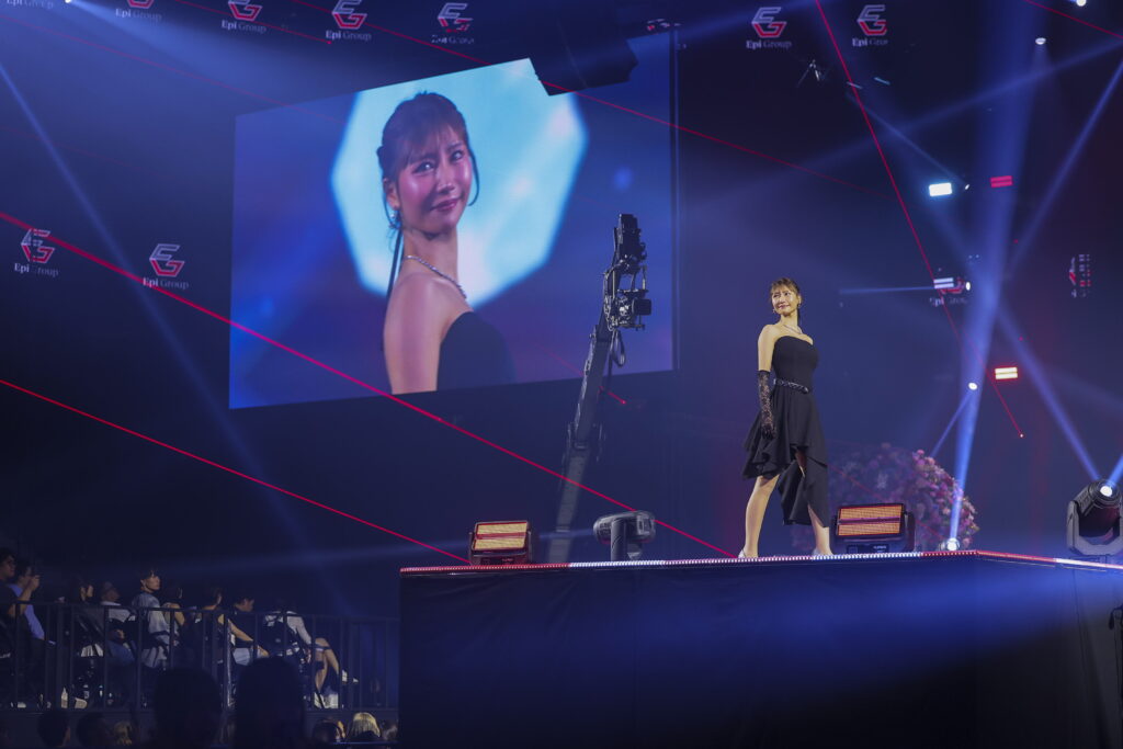 Woman in a black strapless dress stands on a lit stage as a large screen shows her close-up portrait behind her; red laser lines cross the scene with an audience in the foreground.