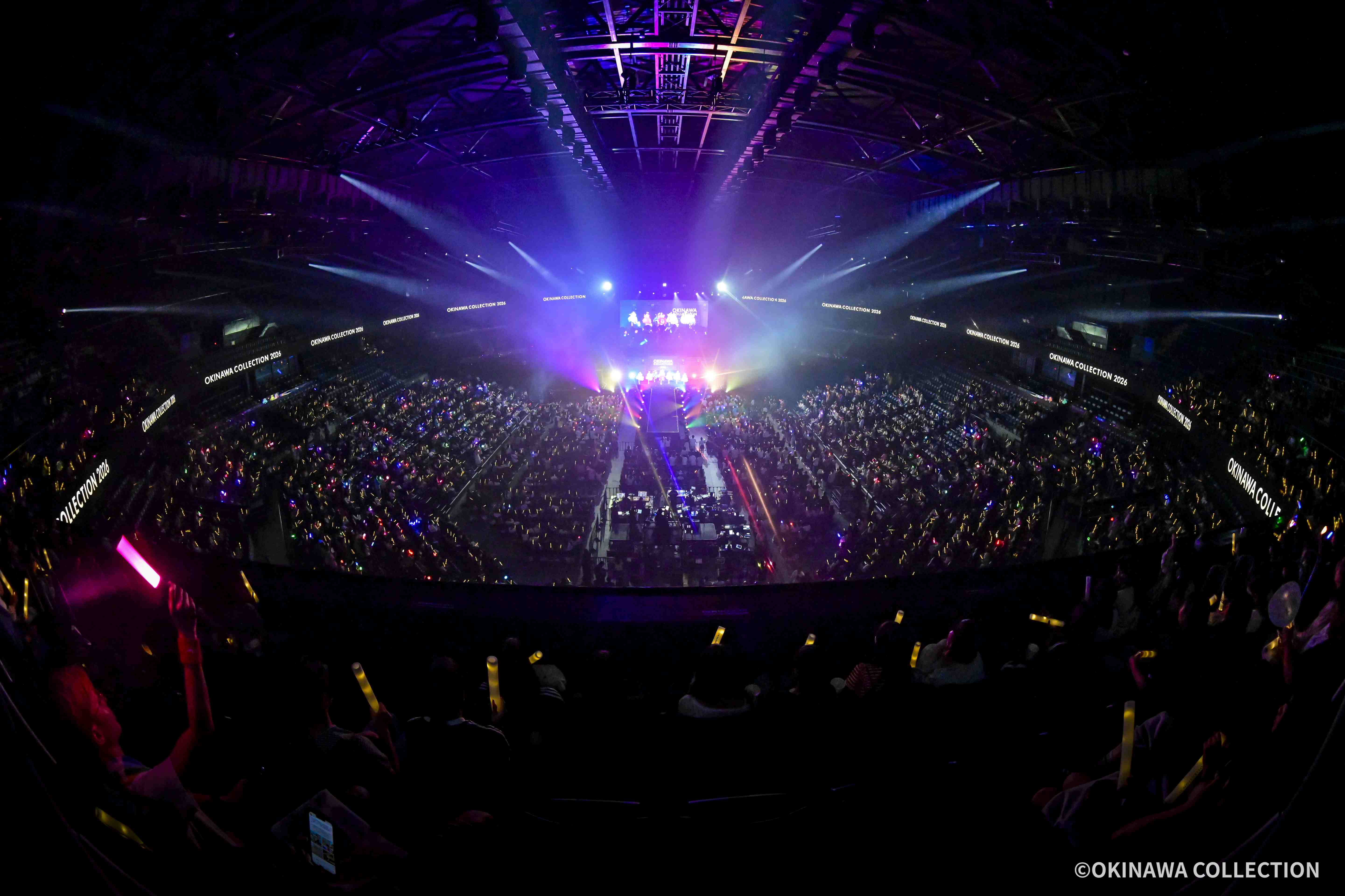 Wide view of a packed arena concert with bright blue and purple stage lights and screens reading 'OKINAWA COLLECTION 2026' along the sides.