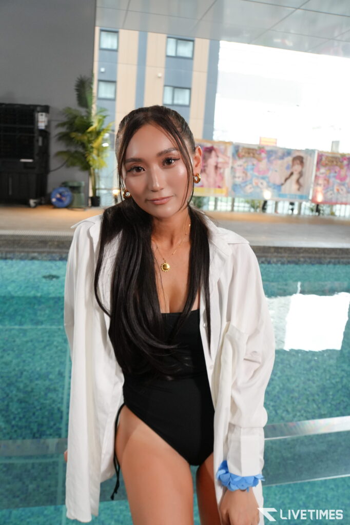 Woman with long black hair in a black swimsuit and white shirt, by a blue pool, wearing a gold pendant and a blue scrunchie, smiling at the camera.