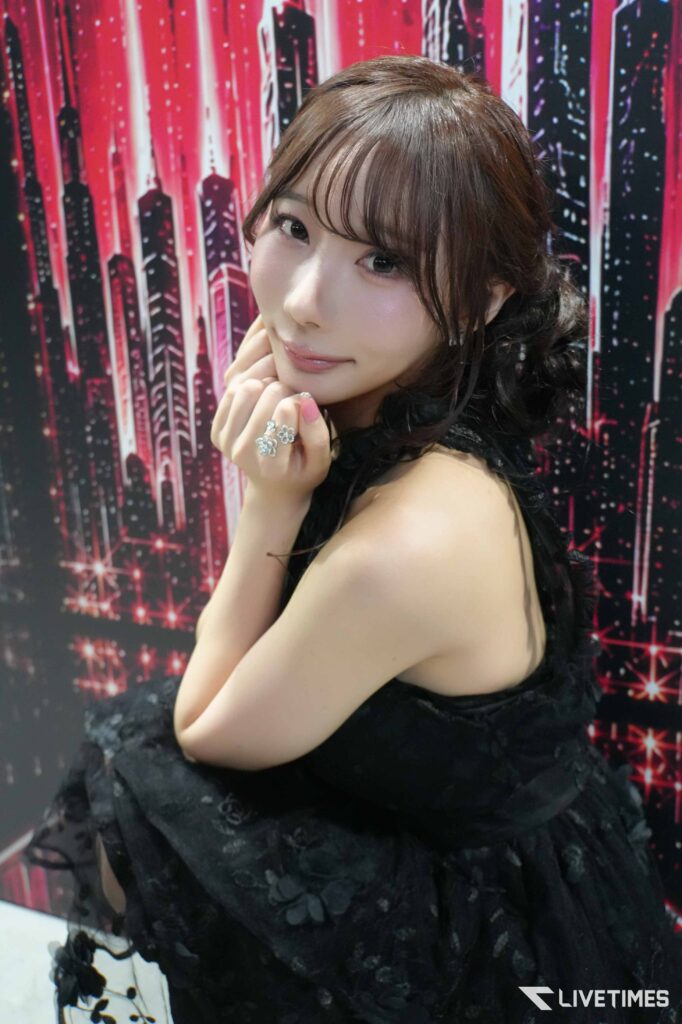 Portrait of a young woman in a black lace dress posing with her hands near her chin against a vibrant pink and black cityscape backdrop.